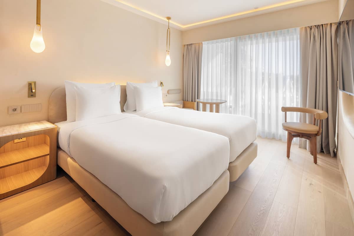A hotel room with two beds, a small table, a chair, and a large window with curtains.