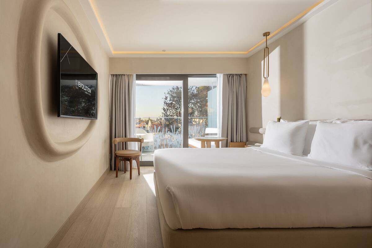 A modern hotel room with a large bed, a flat-screen TV mounted on the wall, a small table, a chair, and a balcony with a view of the city.