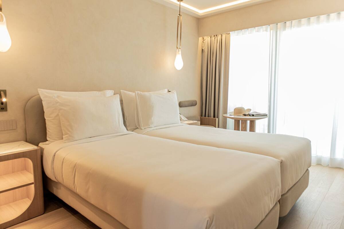 A hotel room with two beds, a bedside table, a lamp, a small table with a hat and sunglasses, and a window with curtains.