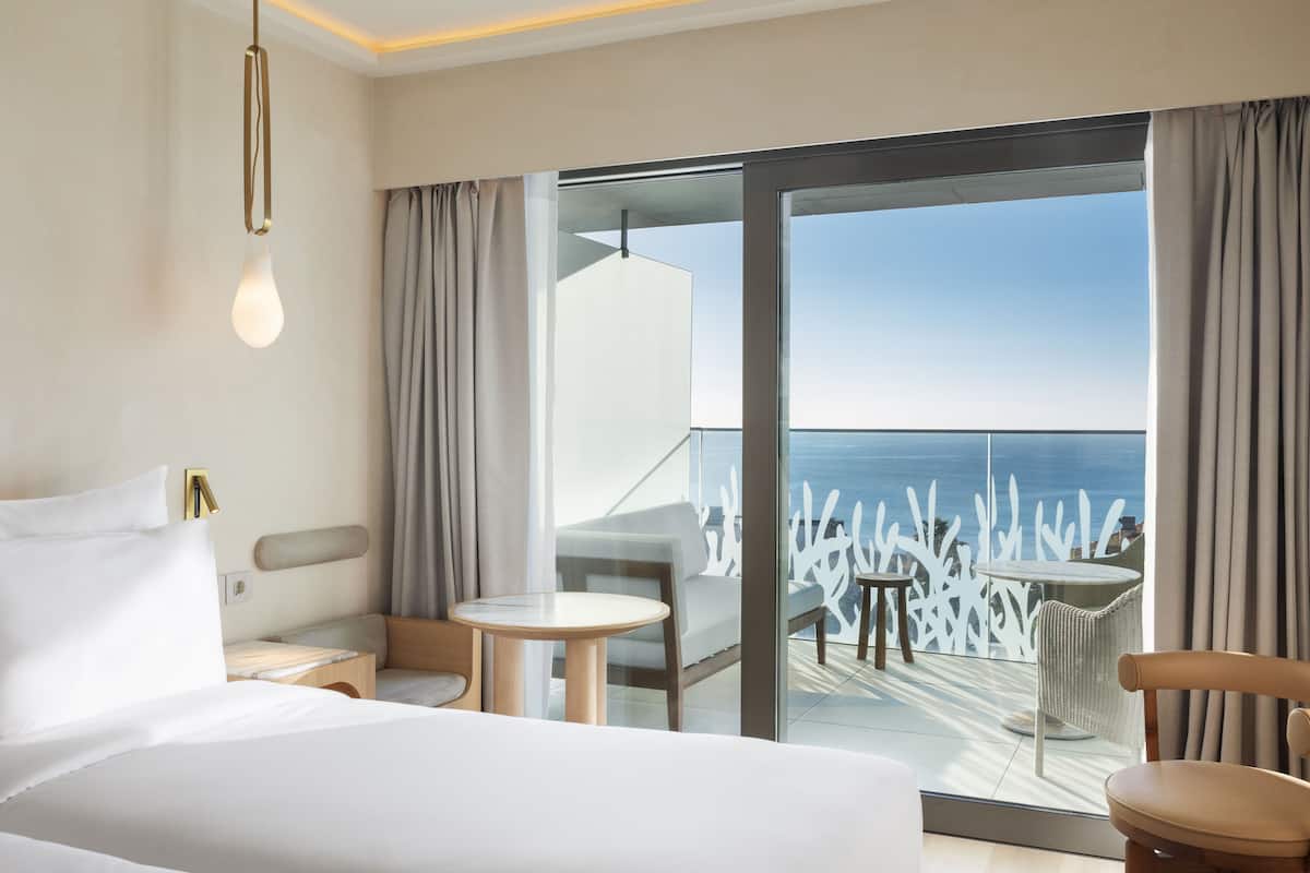 A modern hotel room with a large bed, a small table, and a view of the ocean through a sliding glass door.