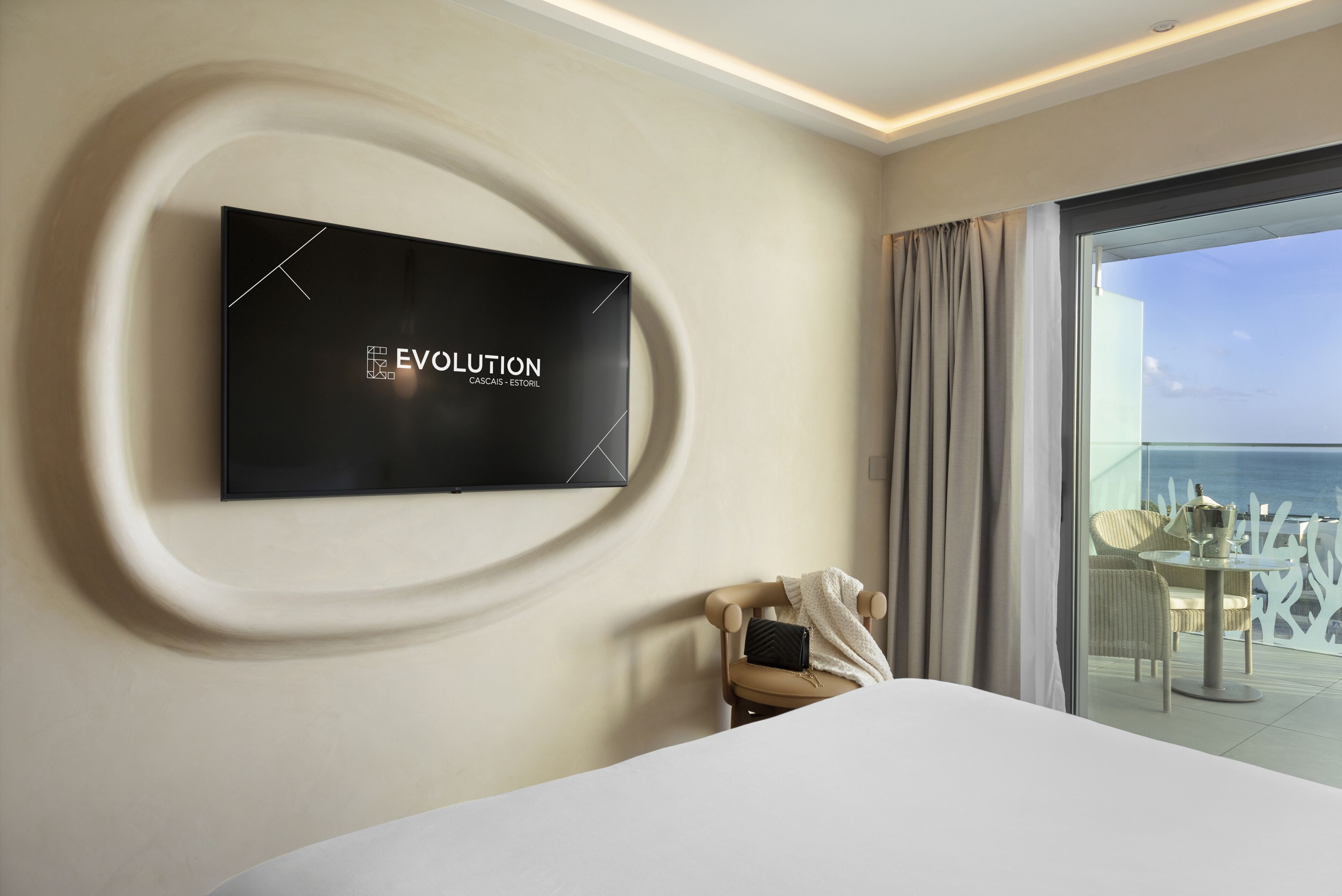 A modern hotel room with a large flat-screen TV mounted on the wall, a bed, a chair, and a balcony with a view of the sea.