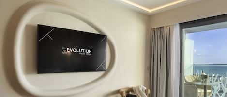 Habitación de hotel moderna con una televisión de pantalla plana grande montada en la pared, una cama, una silla y un balcón con vista al mar.
