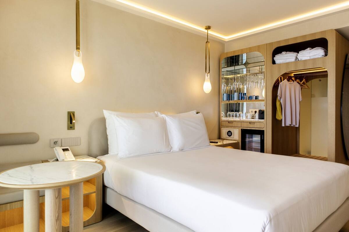 A hotel room with a bed, bedside table, lamp, mirror, wardrobe, and a small table with a phone.