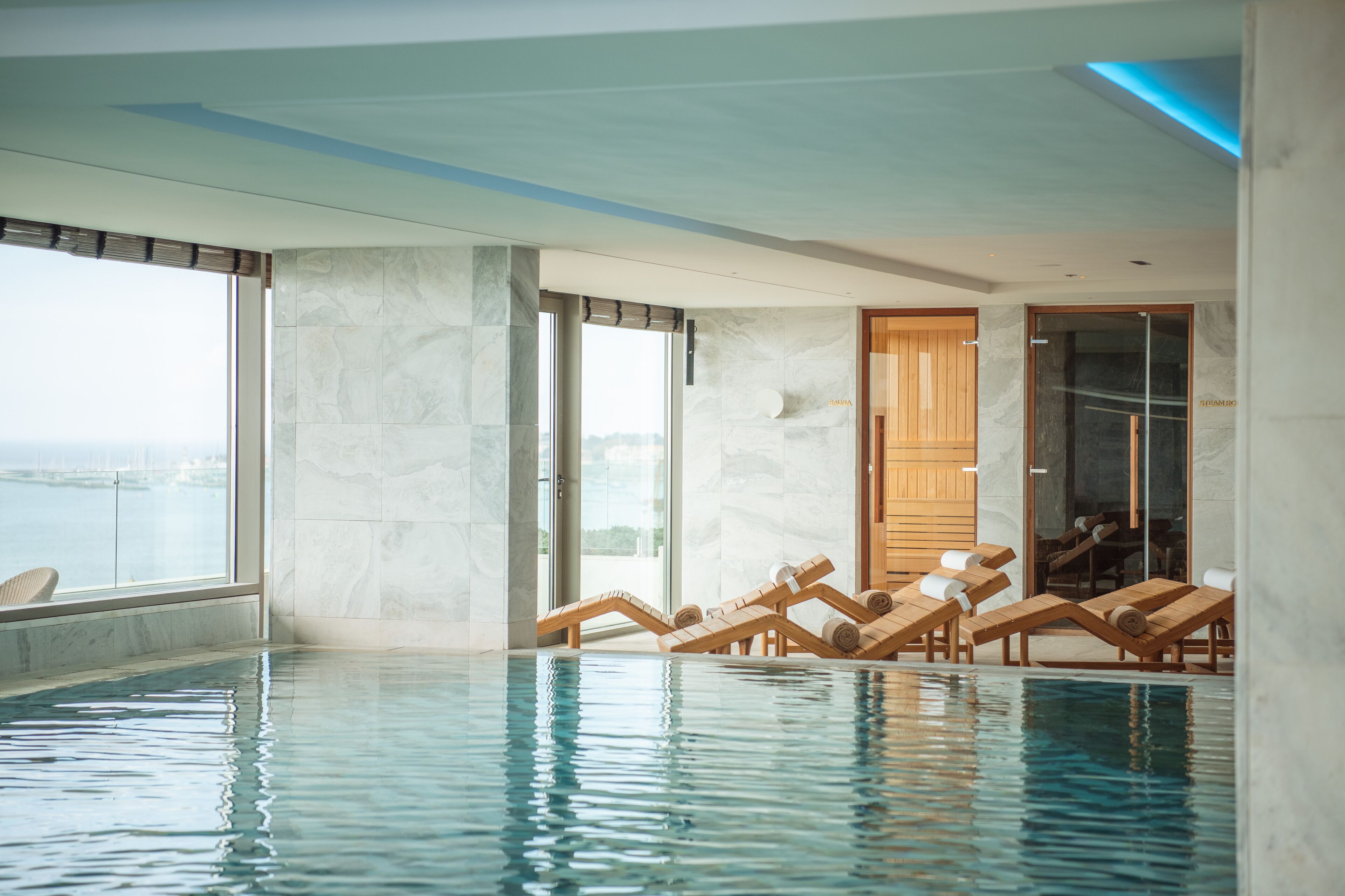 An indoor swimming pool with clear water, wooden loungers, and large windows offering a view of the sea.