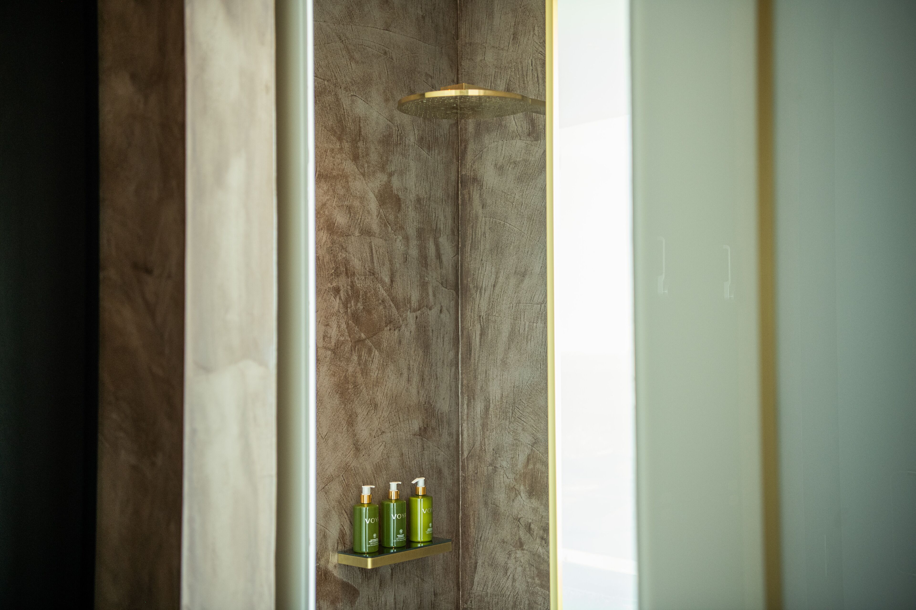 A bathroom with a marble wall, a showerhead, and a shelf with three bottles.