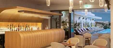 A modern bar area with a curved wooden counter, multiple hanging pendant lights, and wicker chairs arranged around tables.