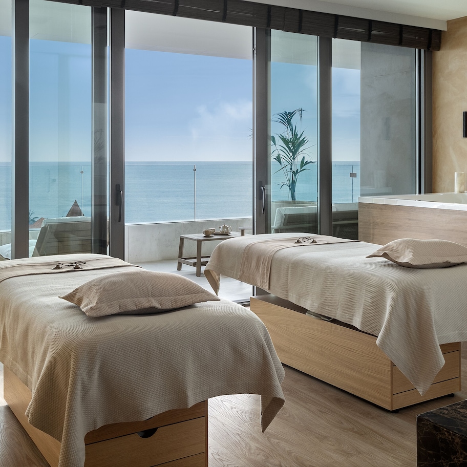 A modern hotel room with two beds, a large window with a view of the sea, a wooden floor, and a marble countertop.