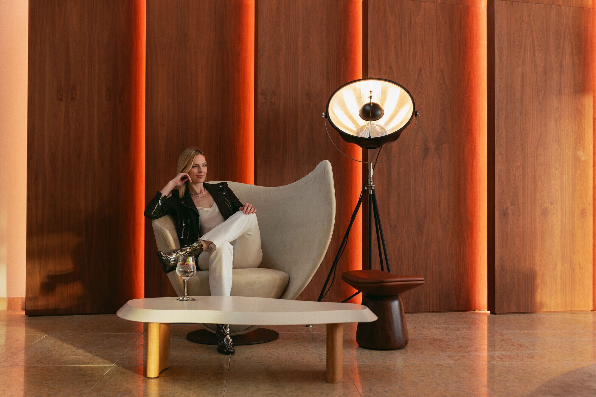 Ein modernes Interieur mit einer Frau, die auf einem gebogenen Sofa sitzt, einem runden Tisch mit einem Weinglas und einer Stehlampe.