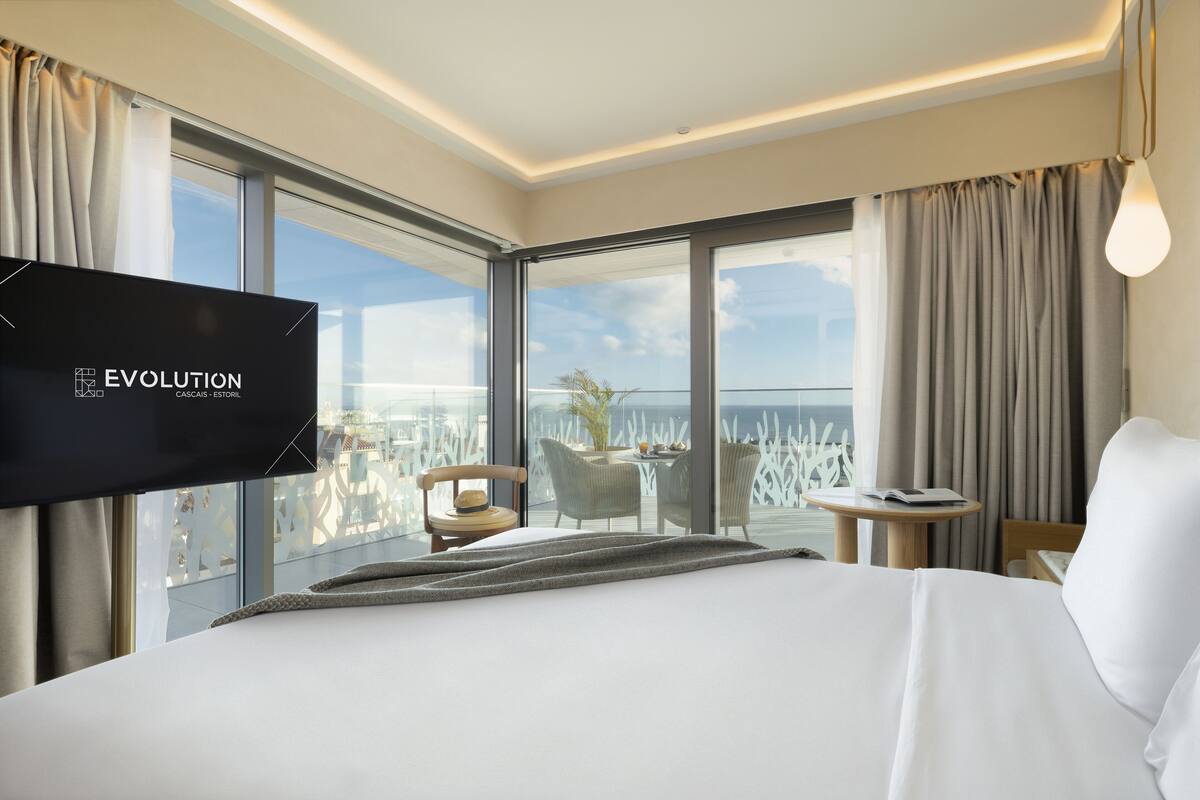 A hotel room with a large bed, a TV, a balcony with a view of the city, and a small table with chairs.