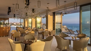Un restaurante con sillas y mesas de mimbre, ventanas amplias con vistas al océano y al paisaje urbano, e iluminación colgante.