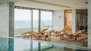 Un área de piscina interior moderna con tumbonas de madera y grandes ventanales con vistas al mar.