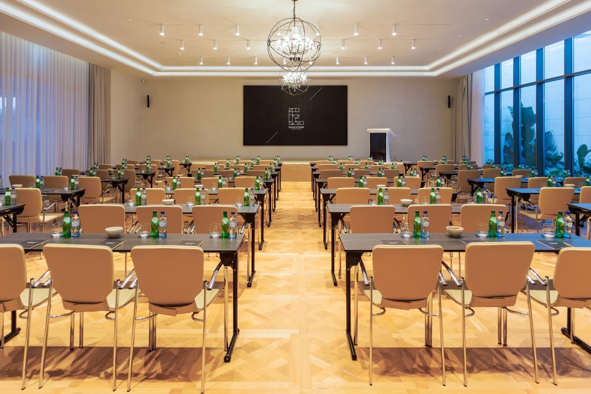 A conference room with rows of tables and chairs, a large screen, and a chandelier.