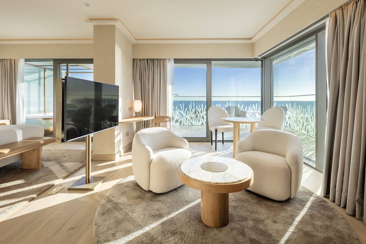 A modern hotel room with a large balcony, plush seating, and a flat-screen TV.