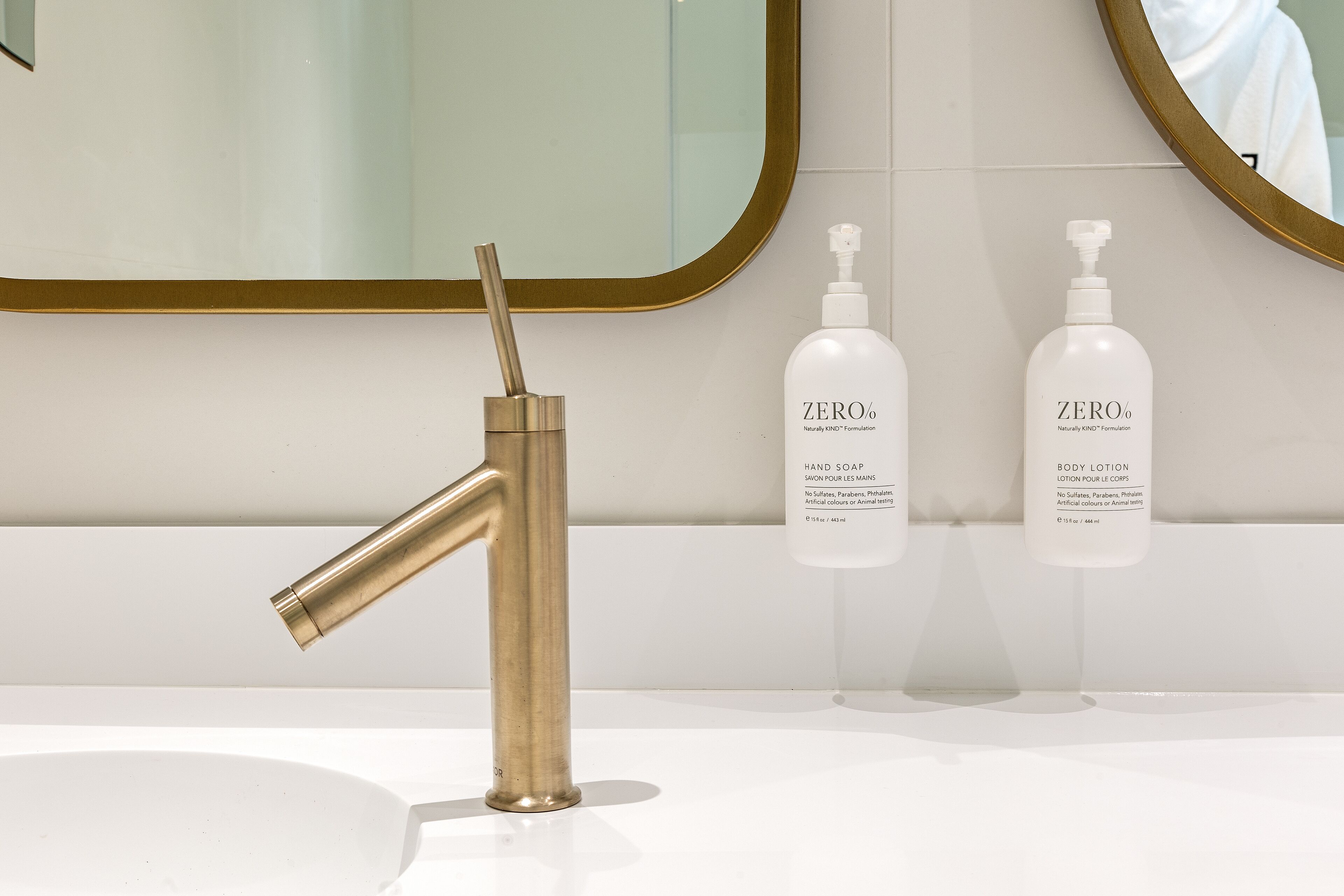 A bathroom with a gold faucet, two bottles of ZERO% hand soap and body lotion, and a mirror with a gold frame.