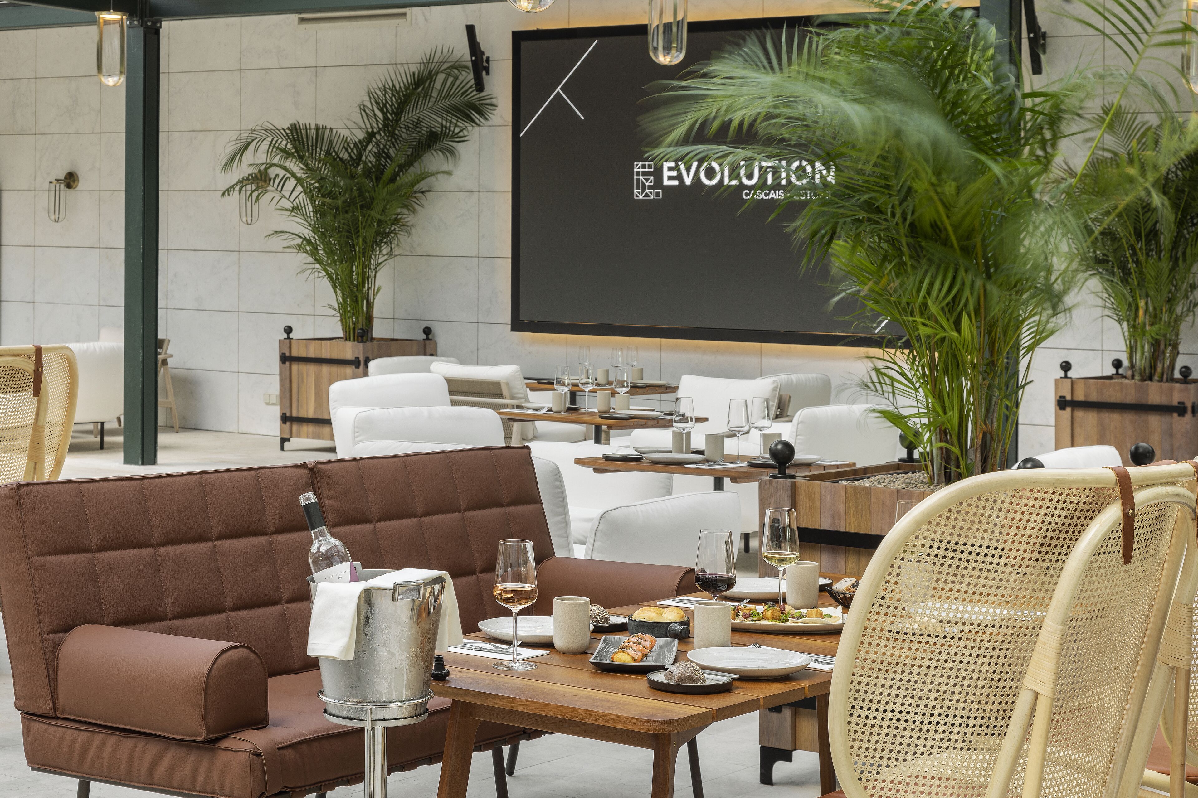 A dining area with a modern interior, featuring a large screen displaying the word "EVOLUTION," a variety of seating options including a brown sofa and wicker chairs, and a buffet table with food and drinks.