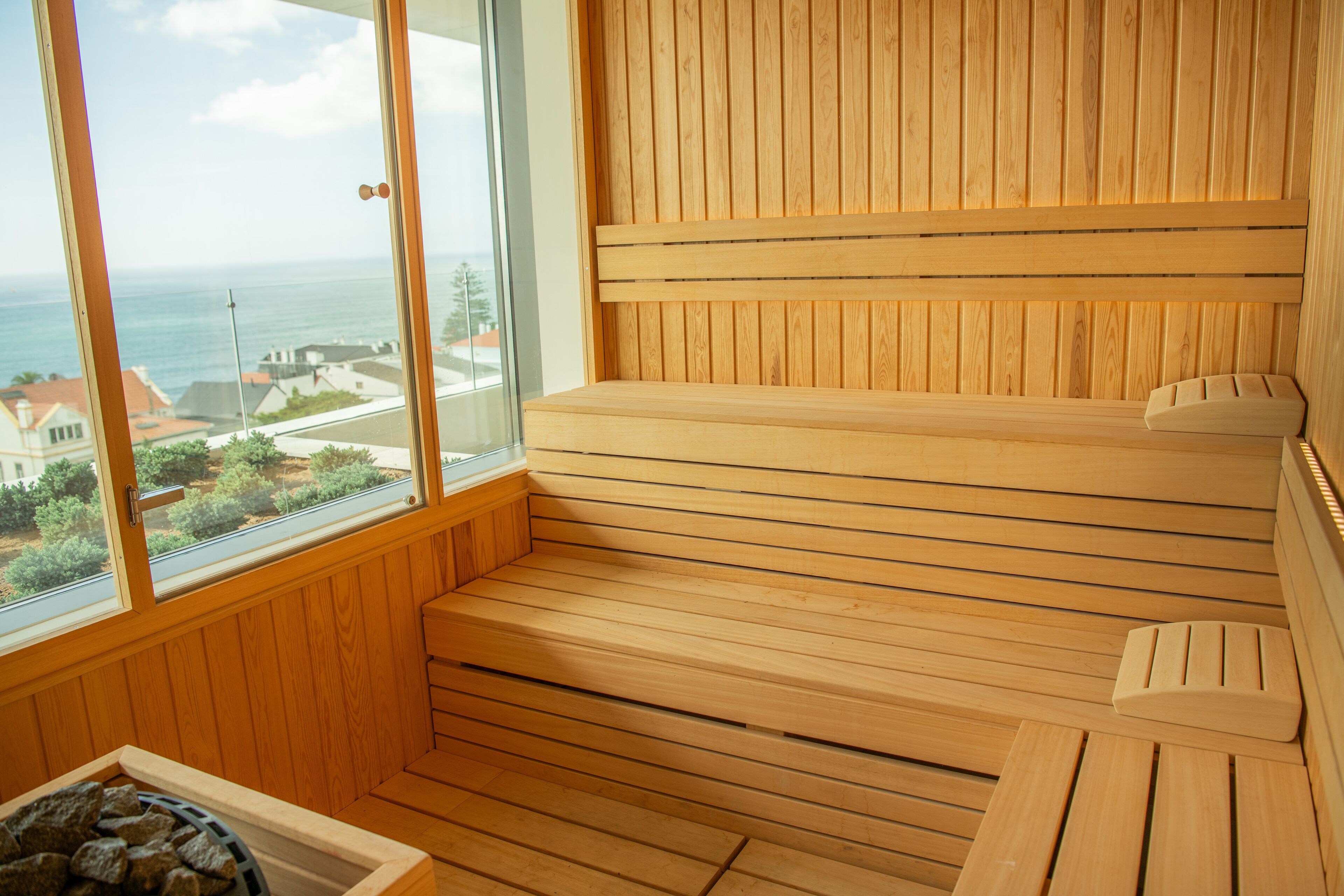 A wooden sauna with a bench and a small stool, a window with a view of the sea and buildings, and a wooden floor.