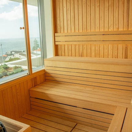 A wooden sauna with a bench and a small stool, a window with a view of the sea and buildings, and a wooden floor.