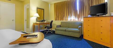Una habitación de hotel con cama, escritorio con lámpara, televisor en una cómoda, sofá y ventana con cortinas.