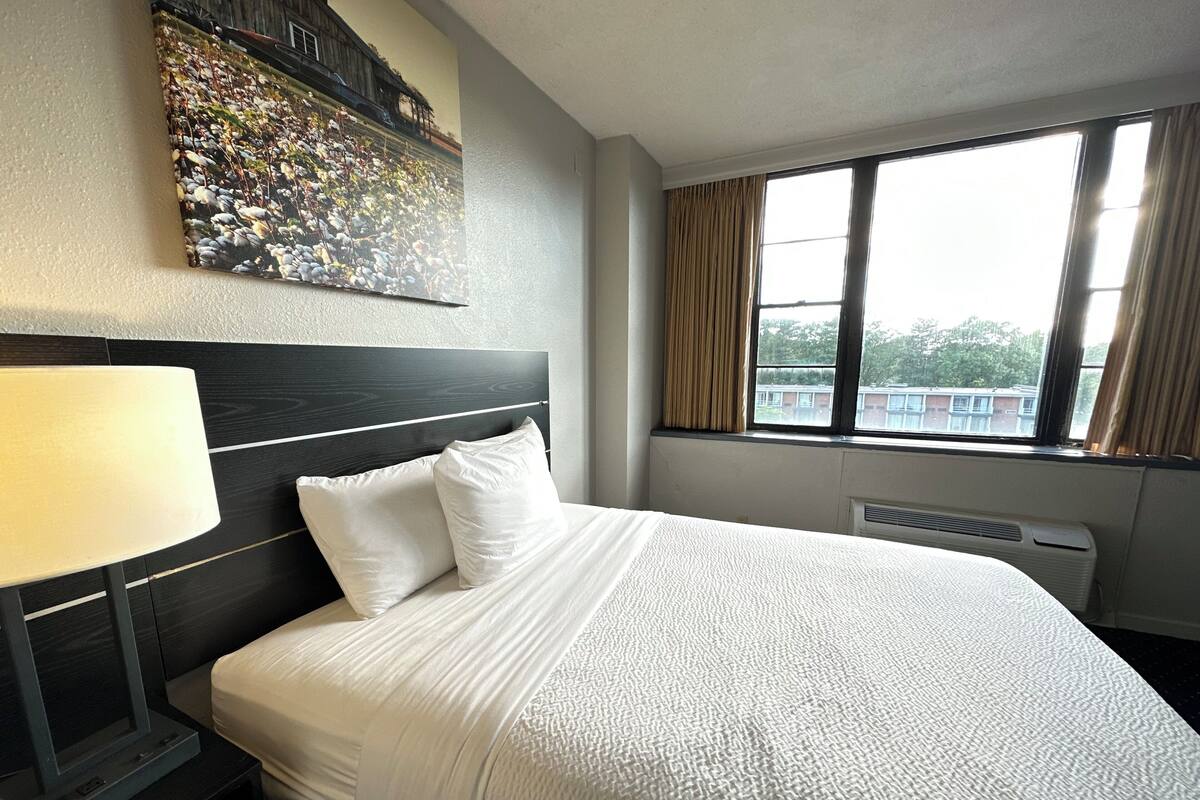 A hotel room with a large bed, a nightstand, a lamp, a window with curtains, and a wall-mounted artwork.