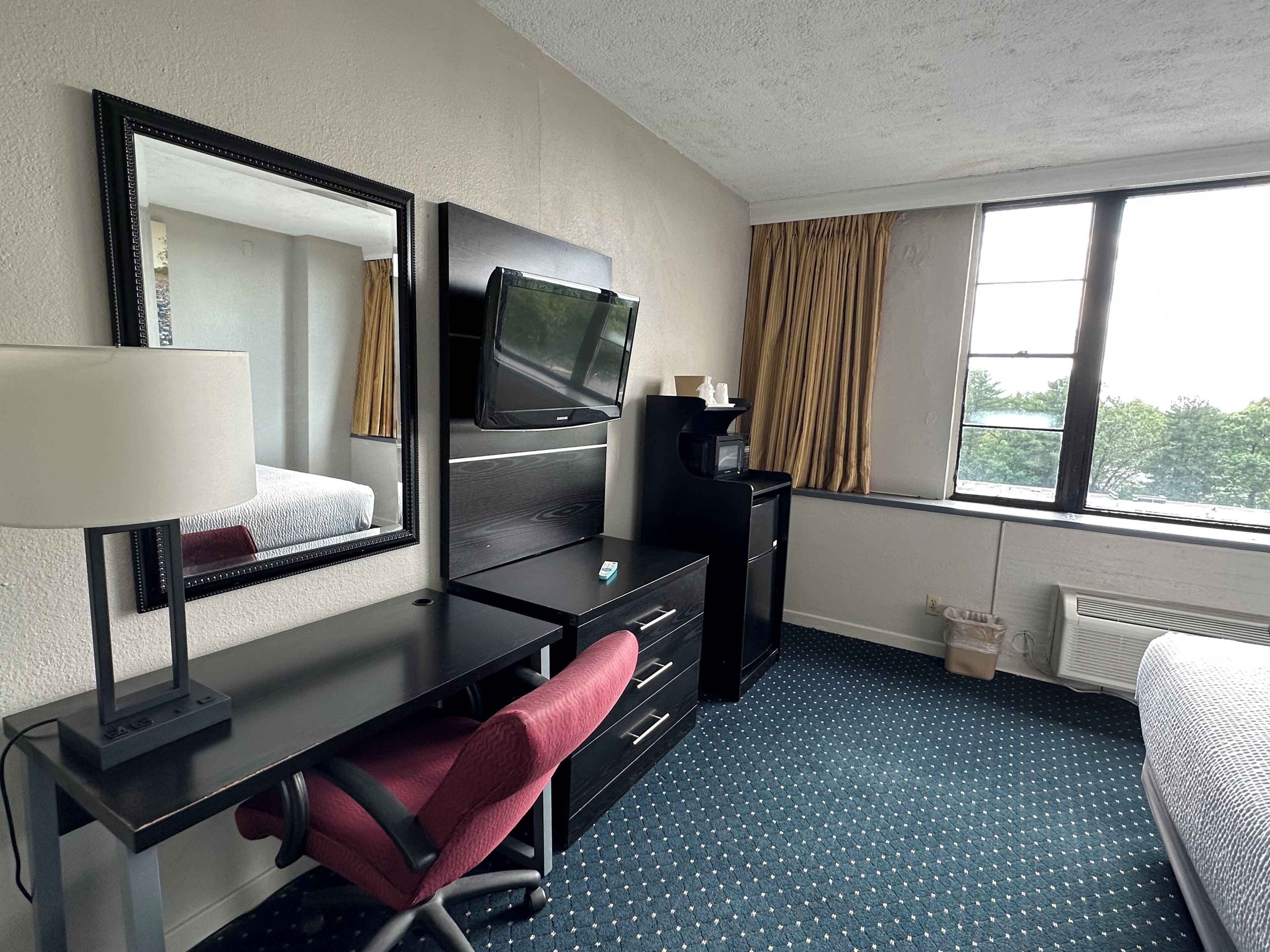 A hotel room with a desk, a TV, a lamp, a chair, and a window with curtains.