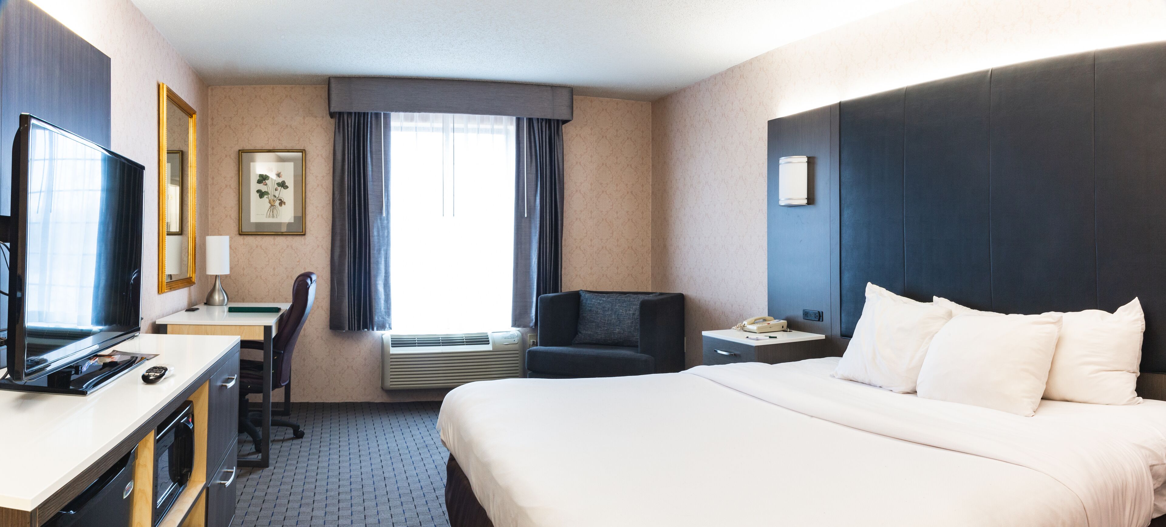 A hotel room with a bed, a desk with a computer, a chair, a TV, a window with curtains, and a nightstand with a phone.
