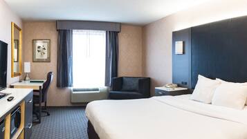 A hotel room with a bed, a desk with a computer, a chair, a TV, a window with curtains, and a nightstand with a phone.