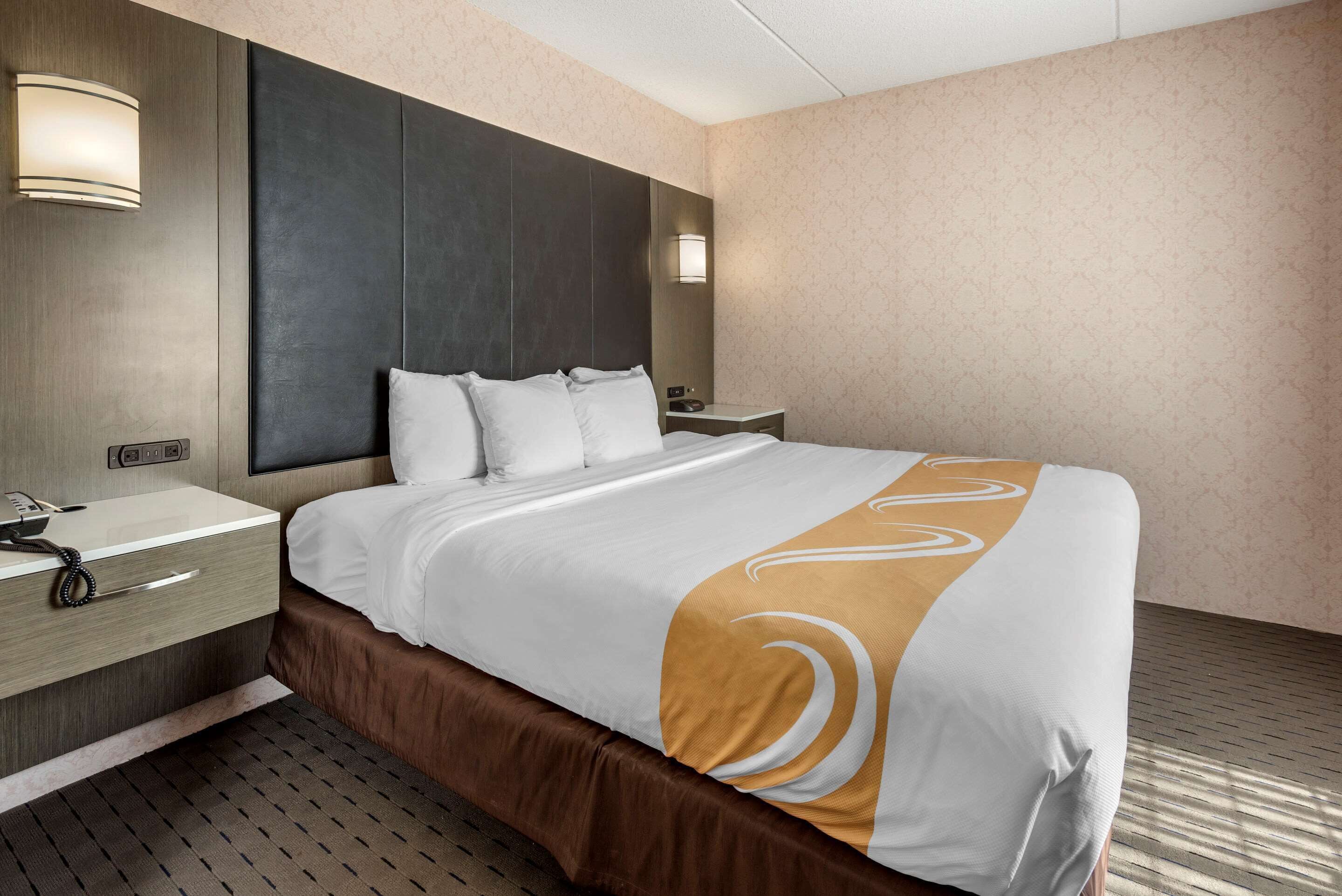 A hotel room with a large bed, a bedside table with a lamp, a telephone, and a wall-mounted light.