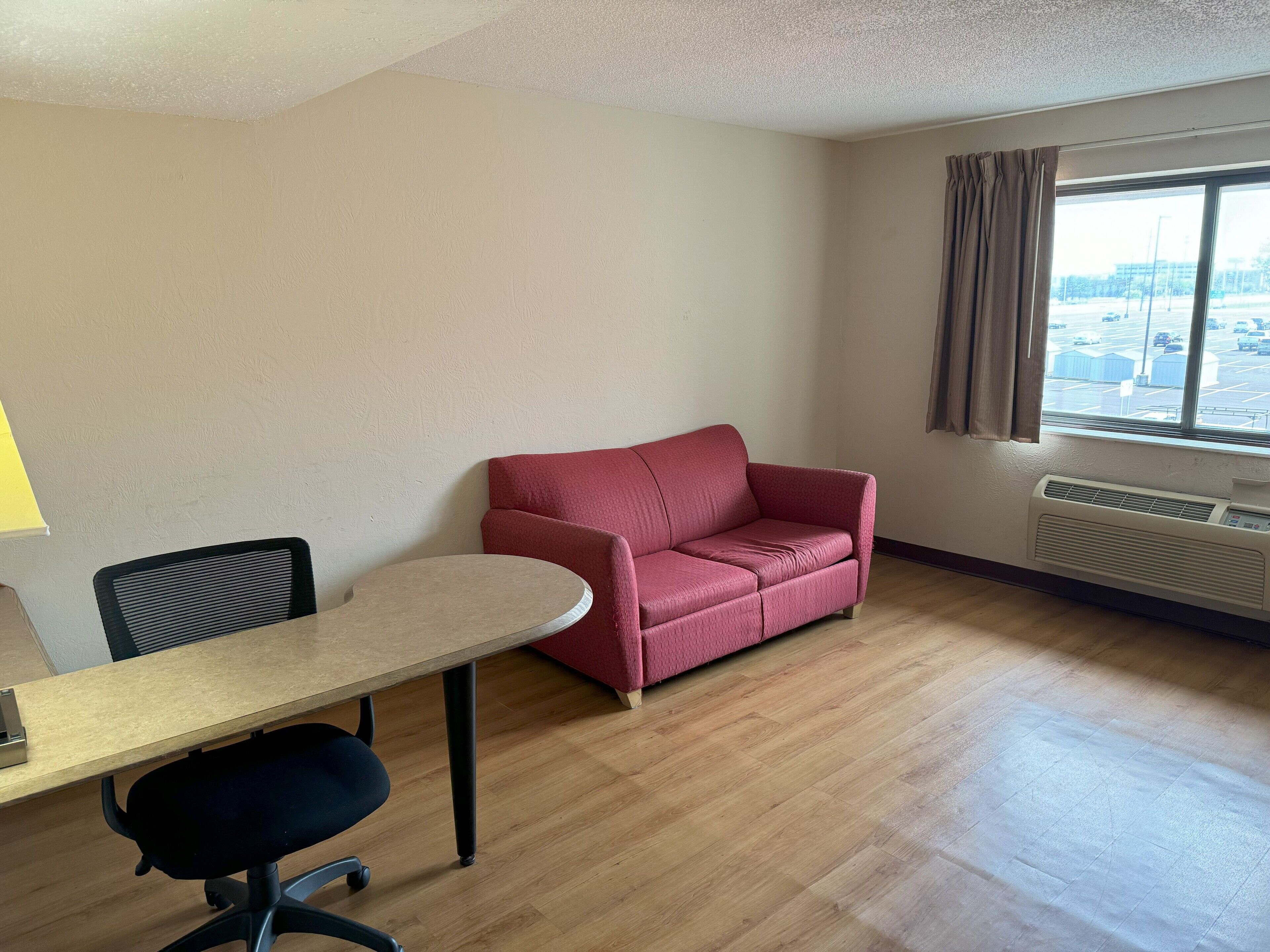 Ein Zimmer mit Holzboden, einem roten Sofa, einem kleinen Tisch mit Stuhl und einem Fenster mit Vorhängen.