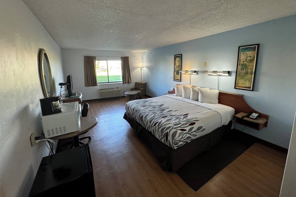 Habitación de hotel con una cama grande, un escritorio con computadora, una silla, una ventana con cortinas, una lámpara y un cuadro en la pared.