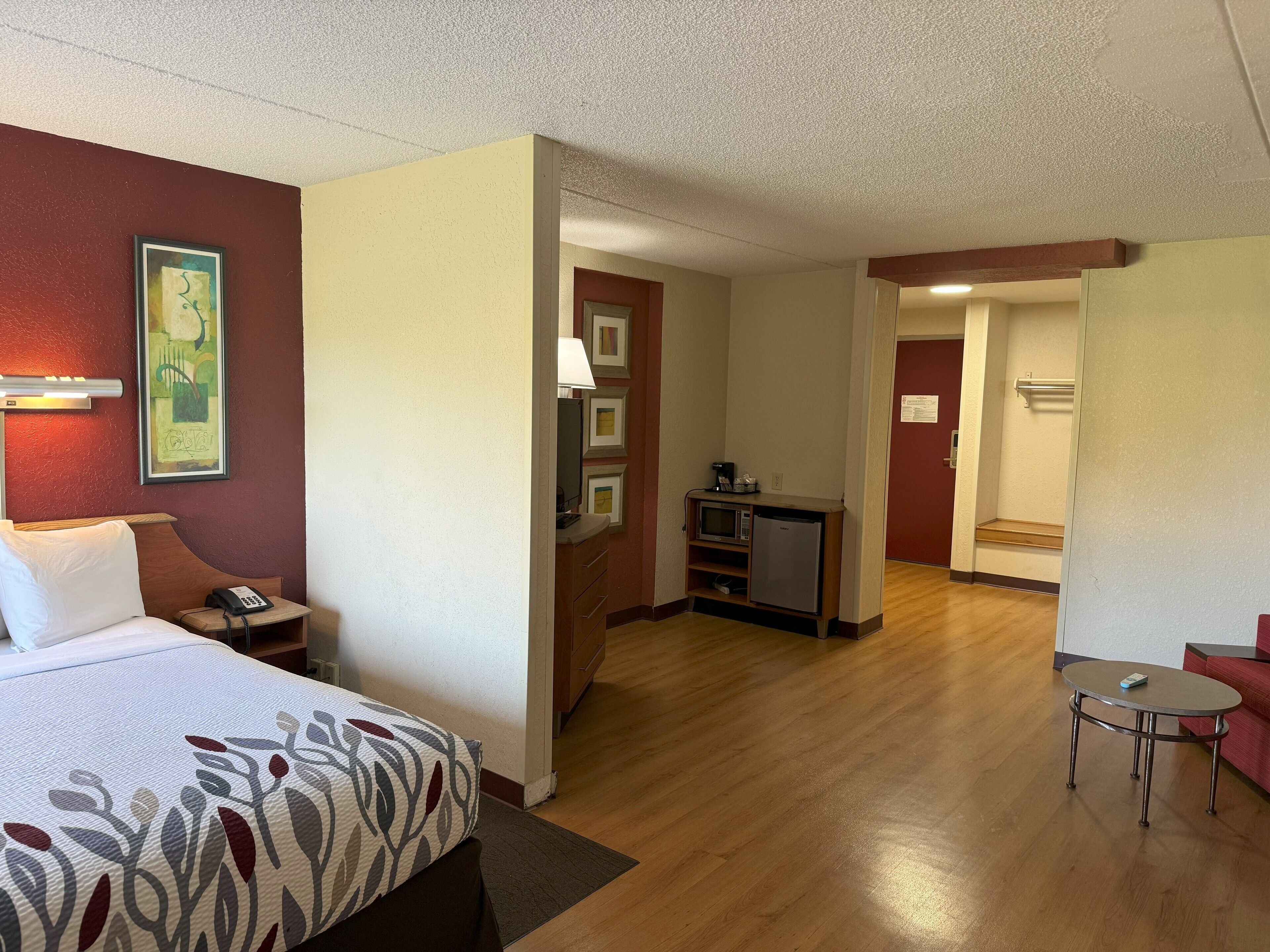 Habitación de hotel con cama, mesita de noche, lámpara y una pequeña kitchenette con microondas y cafetera.