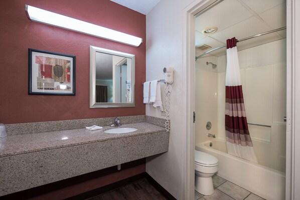 A bathroom with a granite countertop sink, a framed artwork, a hairdryer, and a shower with a striped curtain. - Sycamore Suites (Elyria)