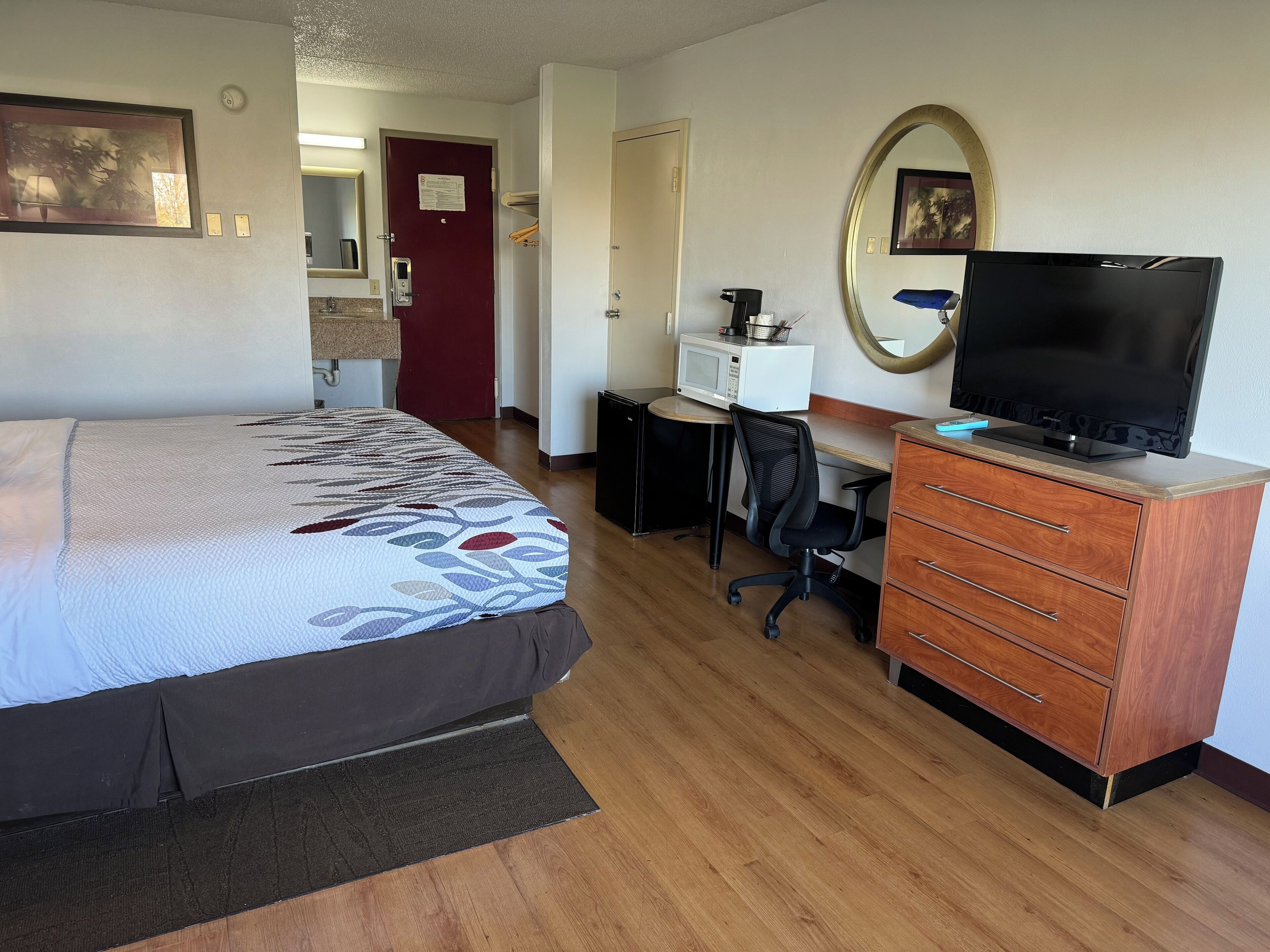 A hotel room with a bed, desk, chair, TV, dresser, microwave, and a mirror.