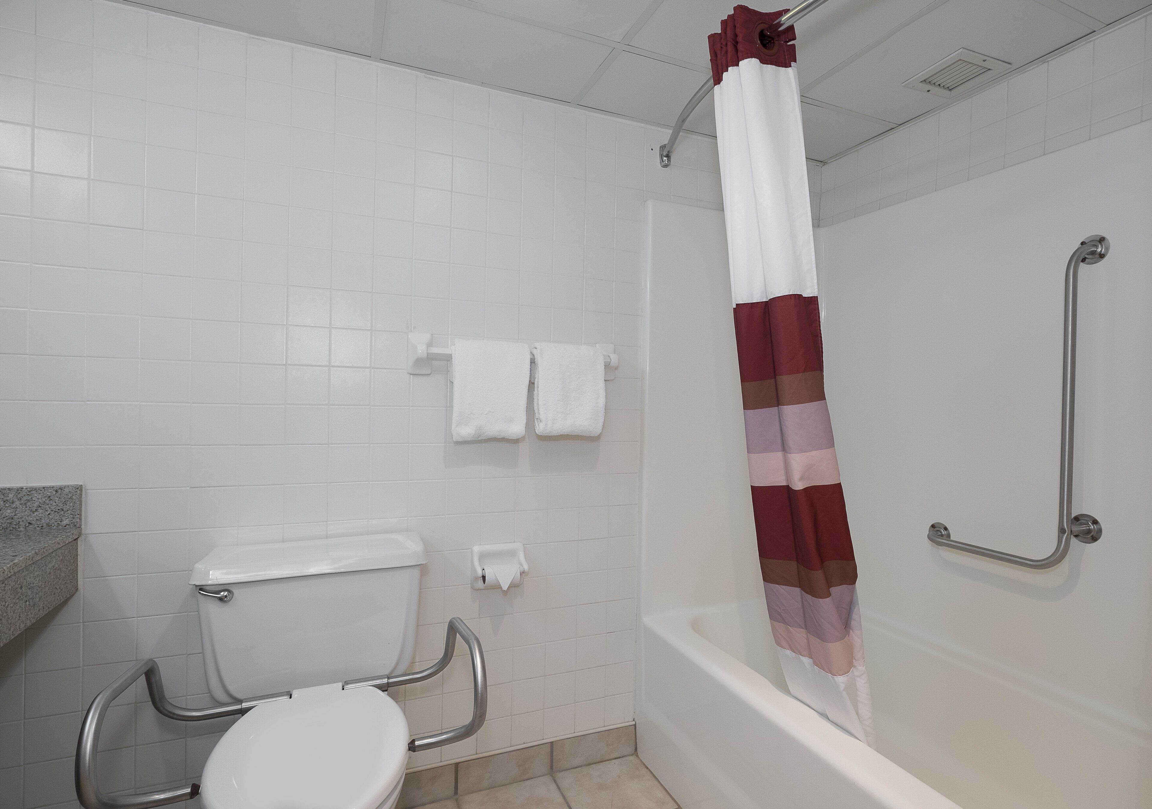 Une salle de bain avec des toilettes blanches, des barres d’appui, une douche avec un rideau à rayures et des murs recouverts de carreaux blancs.