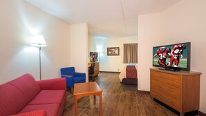 A hotel room with a red sofa, a wooden table, a blue chair, a TV on a wooden cabinet, and a desk with a chair. - Sycamore Suites (Elyria)