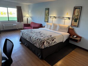 A hotel room with a large bed, a desk with a chair, a window with curtains, and artwork on the walls.