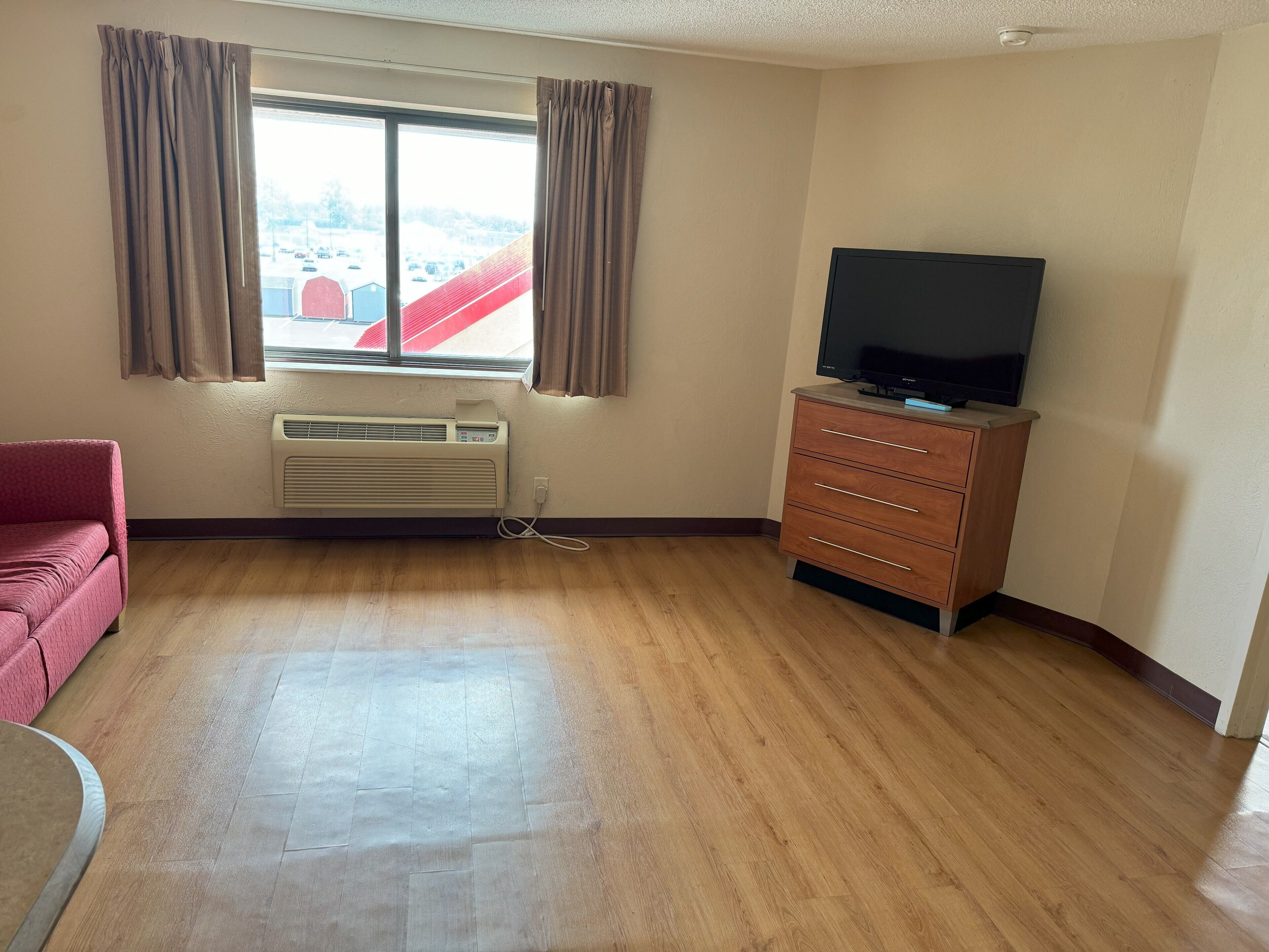 A room with a large window, a flat-screen TV on a wooden cabinet, a red couch, and a wooden floor.