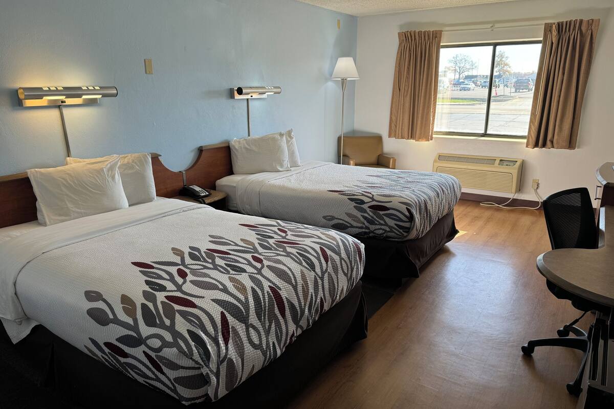 Una habitación de hotel con dos camas, un escritorio, una silla, una lámpara y una ventana con cortinas.
