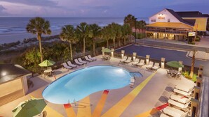 A swimming pool with lounge chairs and umbrellas, a beach view, and a building with a sign.
