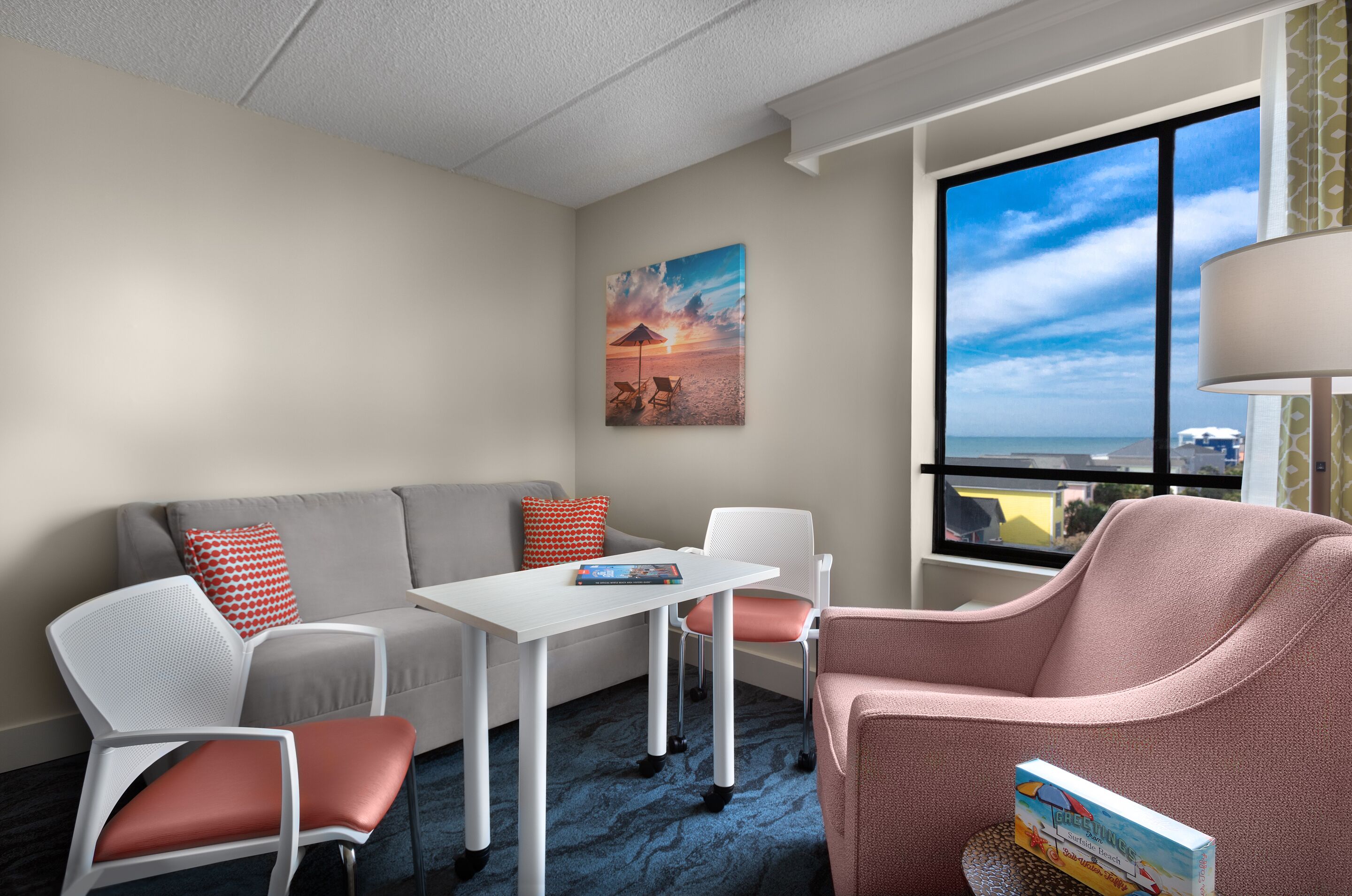 A modern hotel room with a grey sofa, white chairs, a small table, and a large window offering a view of the city.