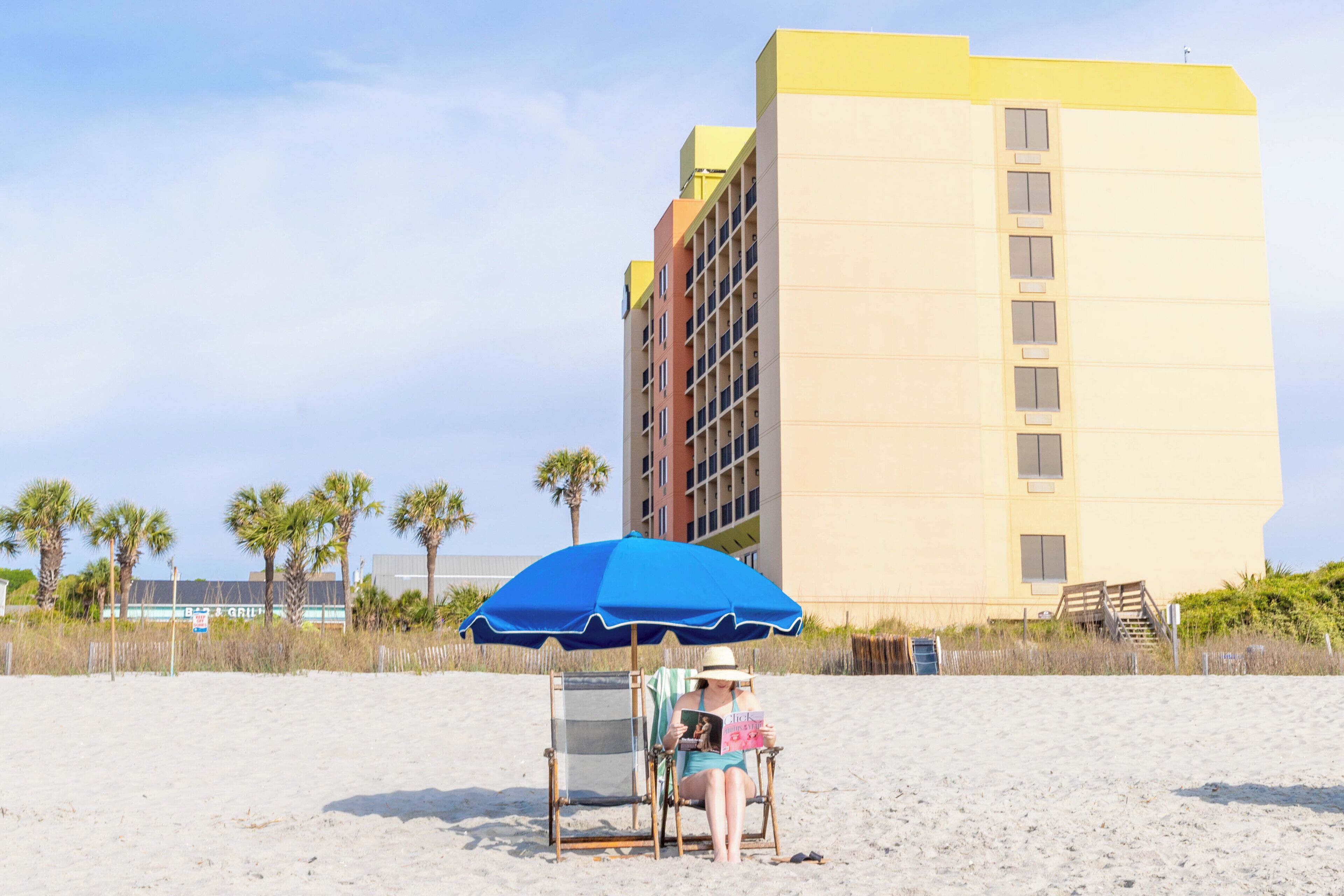 A beach chair with a blue umbrella, a person reading a book, and a building in the background.