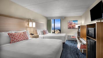A hotel room with two beds, a desk, a TV, and a view of the ocean.