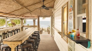 A beachside restaurant with a wooden table and wicker chairs, a menu board, and a view of the ocean.