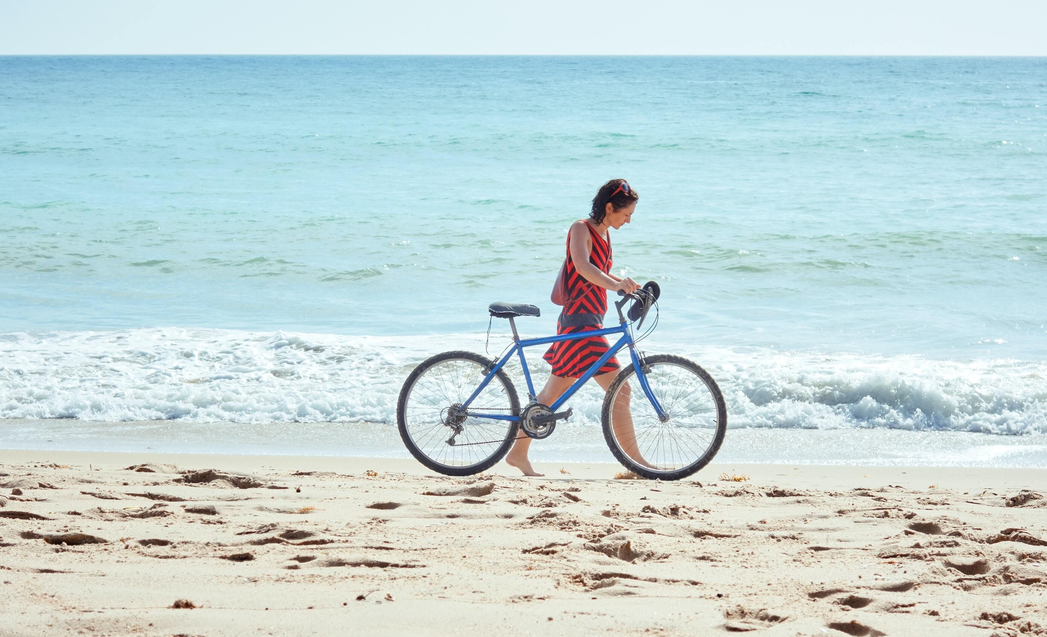 Une personne qui pousse un vélo sur une plage de sable, avec l’océan en arrière-plan.