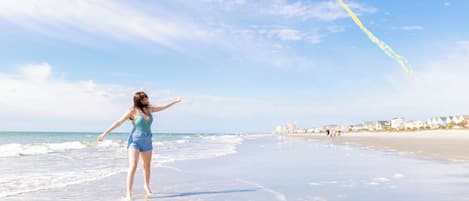 A person flying a yellow kite on a beach with buildings in the background.