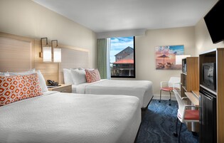 A hotel room with two beds, a desk, a television, and a view of a building outside.
