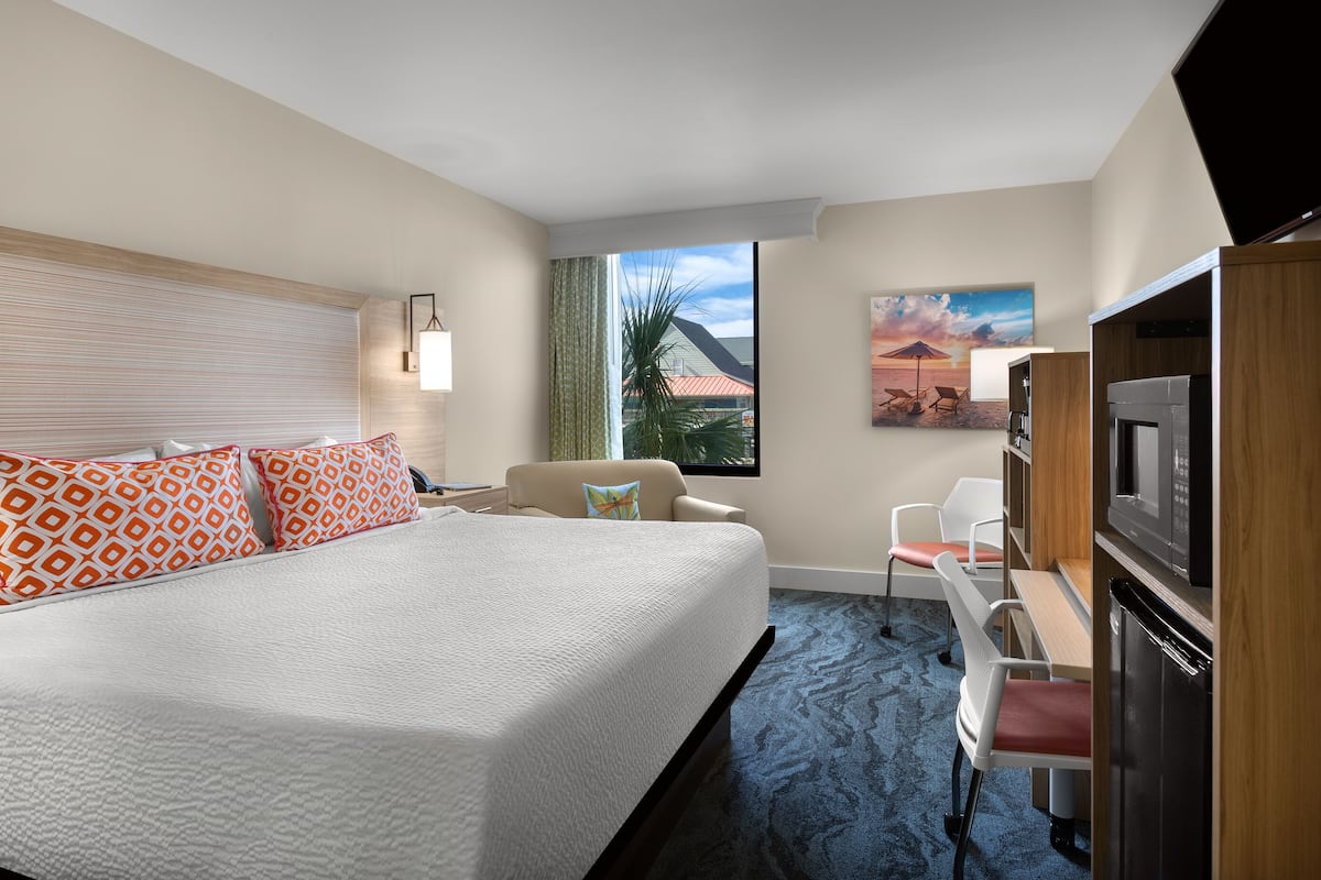 Une chambre d’hôtel avec un grand lit, un bureau avec une chaise, une télévision et une fenêtre donnant sur des palmiers et un coucher de soleil.