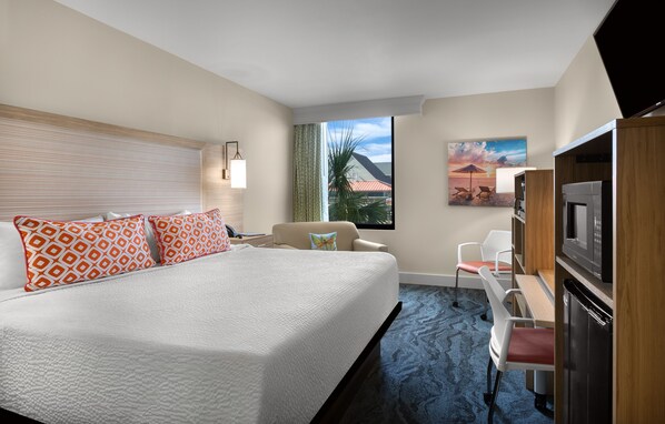 Habitación de hotel con una cama grande, un escritorio con silla, televisión y ventana con vista a palmeras y un atardecer.