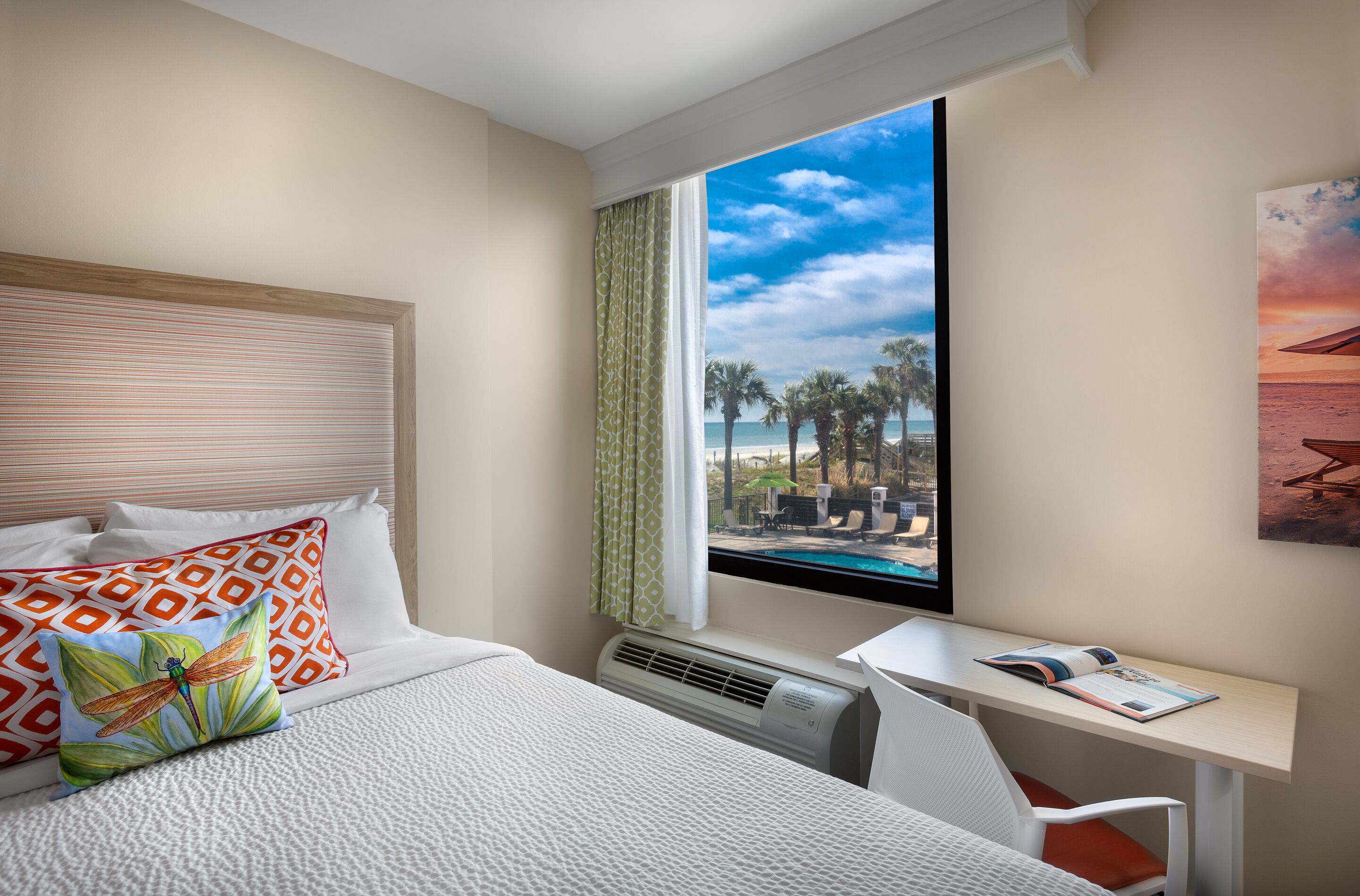 A hotel room with a bed, a desk, a window with a view of the beach, and a mounted air conditioner.