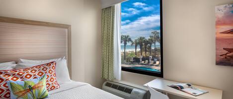 A hotel room with a bed, a desk, a window with a view of the beach, and a mounted air conditioner.