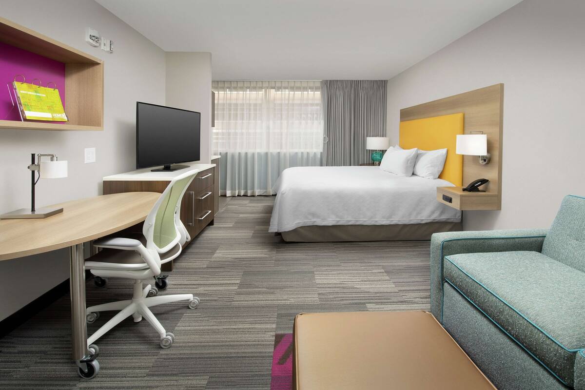 A hotel room with a bed, a desk with a computer, a TV, a sofa, and a bedside table with a lamp.